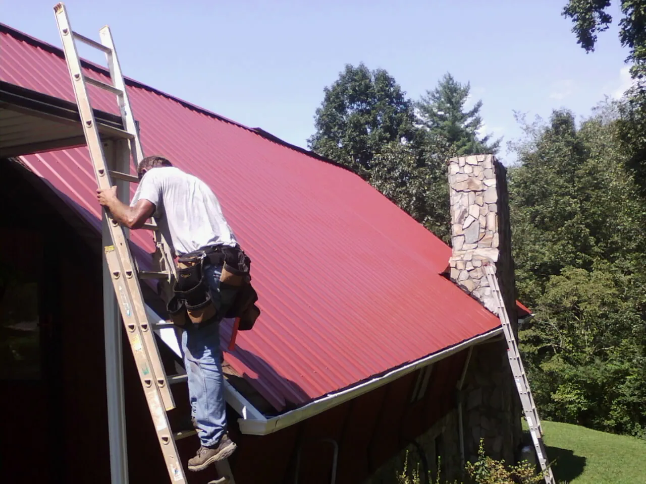 Roofer on a ladder beside a red metal roof and stone chimney — Soffit & Fascia Repair by Roofing Brothers in Red Lodge